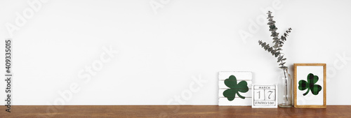 St Patricks Day decor on a wood shelf. Shabby chic wood signs, calendar and green branches against a white wall banner. Copy space.
