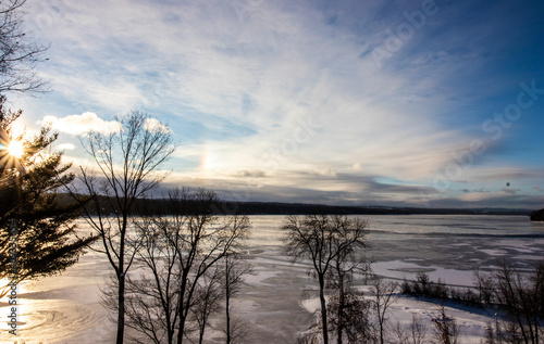 Frozen lake in winter with ice in Canada  dramatic sky and sunlight reflecting in the lake  bare trees in foreground  natural sunlight  horizontal image