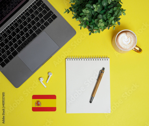 online Spanish language course moke up. Layout of laptop, headphones, Spanish flag, notepad with pen, cappuccino coffee and plant on yellow background