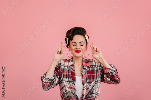 Pinup girl posing with eyes closed and crossed fingers. Cheerful caucasian lady in checkered shirt standing on pink background.