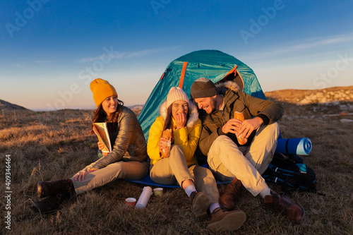 A group of young friends sitting and eat sandwiches, enjoying the mountain sun next to the tent.