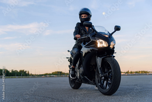 Attractive girl in black leather jacket, pants and helmet on outdoors parking rides on stylish sports motorcycle at sunset.