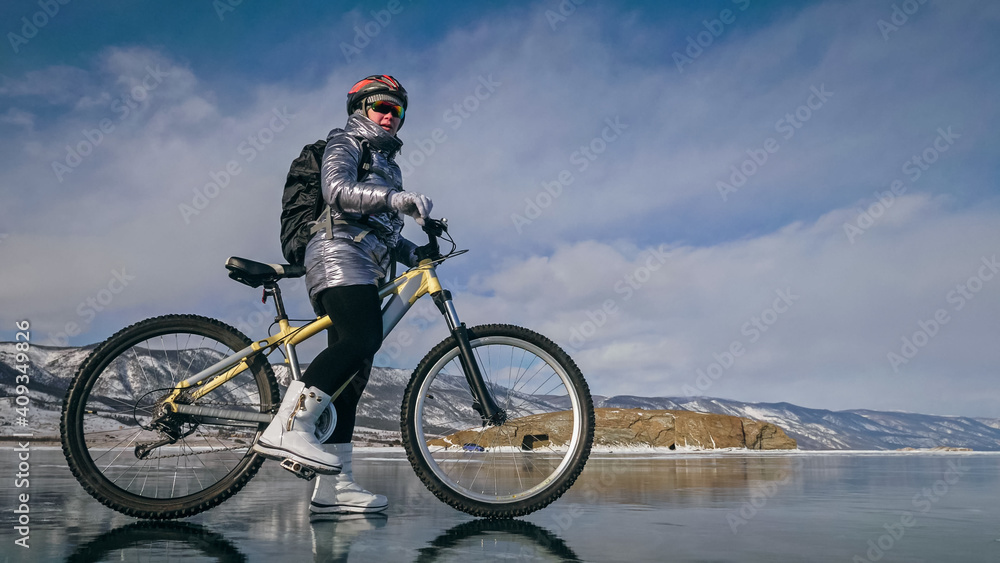 Woman is riding bicycle on the ice. Girl is dressed in a silvery down jacket, cycling backpack and helmet. Ice of the frozen Lake Baikal. Tires on bike are covered with spikes. Traveler is ride cycle.