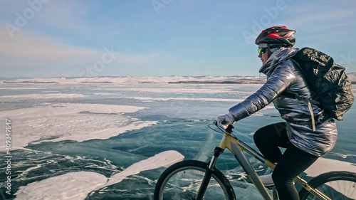 Woman is riding bicycle on the ice. Girl is dressed in a silvery down jacket, cycling backpack and helmet. Ice of the frozen Lake Baikal. Tires on bike are covered with spikes. Traveler is ride cycle. photo