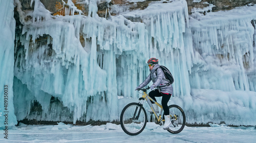 Woman is riding bicycle near the ice grotto. The rock with ice caves and icicles. Girl is dressed in silvery down jacket, cycling backpack and helmet. Tires winter with spikes. Traveler is ride cycle. photo