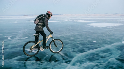 Fototapeta Naklejka Na Ścianę i Meble -  Man is riding bicycle on the ice. Ice of the frozen Lake Baikal. Teenage is dressed in black down jacket, cycling backpack, helmet. Tires on covered with special spikes. Traveler boy is ride cycle.