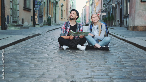 Trendy couple of tourists using map, sitting on pavement and admiring historical surroundings. They are excited and smiled. © art24pro