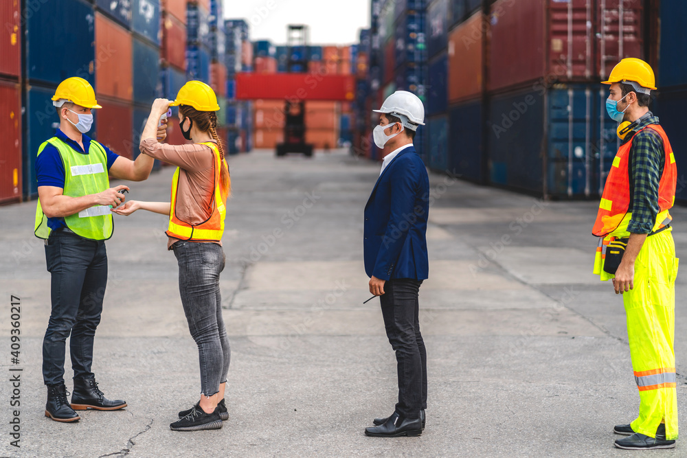 Professional of group cargo foreman in helmets standing and using infrared thermometer for checking body temperature staff fever before work in quarantine for coronavirus wearing protective mask Professional of group cargo foreman in helmets standing and using infrared thermometer for checking body temperature staff fever before work in quarantine for coronavirus wearing protective mask