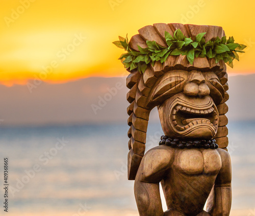 Hawaiian Tiki Statue with an amazing sunrise in the background. The tiki is dressed up with traditional leaves and a macadamia necklace. It is made of wood and looks large in size.