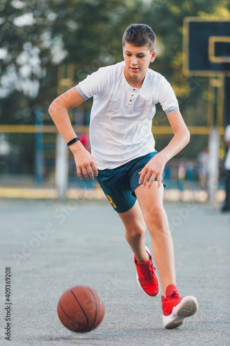 Cute young boy plays basketball on street playground in summer. Teenager in white t shirt with orange basketball ball outside. Hobby, active lifestyle, sport activity for kids. © Natali
