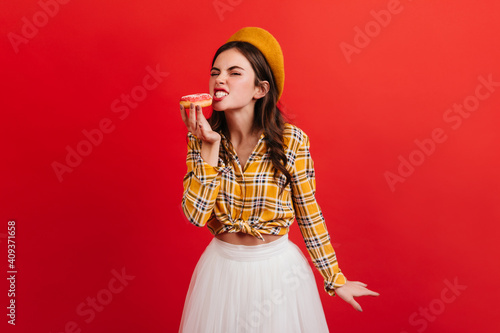 Hungry girl in stylish outfit bites strawberry donut. Portrait of woman in checkered blouse and orange beret on bright background