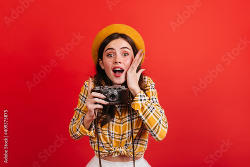 Beautiful young girl blogger makes photo on retro camera. Portrait of green-eyed woman in orange outfit and hat on red background