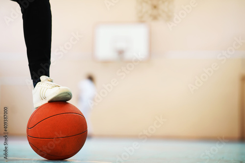 A human foot rest on the basketball on the concrete floor. Photo of one basket ball and sneakers in a wooden floor.