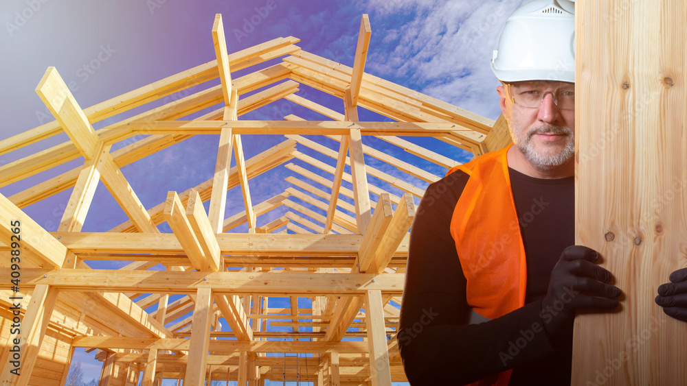 Fototapeta premium The Builder holds up the boards and smiles at the camera. A man in a construction helmet on the background of a wooden frame of a cottage. High-quality lumber. Construction of country houses.