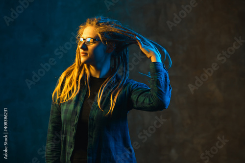 Young woman with dreadlocks and glasses in yellow and blue neon lighting on dark background. Stylish girl in futuristic environment.