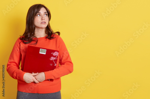 Obraz na plátne Diet and weight, young frustrated woman, looking aside with pensive facial expression, feeling unhealthy, embracing mechanical scales, wearing orange sweater, poses isolated over yellow background