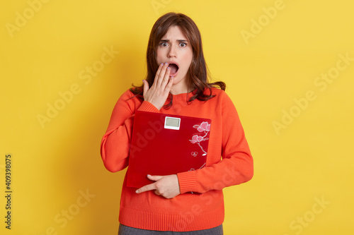Photos Frustrated woman with scale in hands posing isolated over yellow background, female being very shocked, cover opened mouth with palm, needs to keep fit and loosing weight