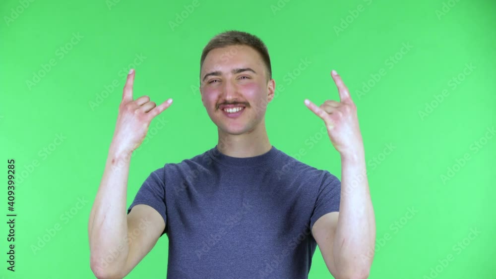 Portrait of a young man making a rock gesture, enjoying life and laughing. Cute male with a mustache in a blue t-shirt posing on a green screen in the studio. Close up. Portrait of a young man making a rock gesture, enjoying life and laughing. Cute male with a mustache in a blue t-shirt posing on a green screen in the studio. Close up.