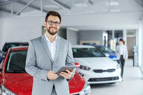 Smiling friendly car seller in suit standing in car salon and holding tablet. It's always pleasure to buy a car on a right place. photo