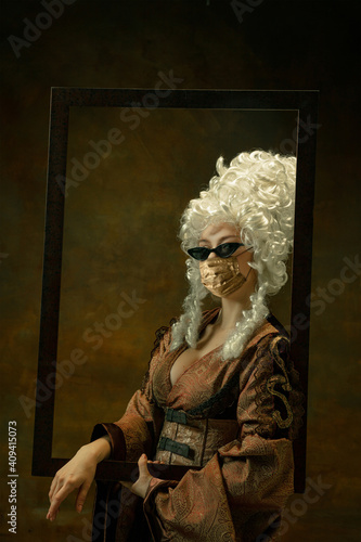 Portrait of medieval young woman in vintage clothes, golden face mask with picture frame on dark background. Royal person protected from covid. Concept of comparison of eras, modern, fashion.