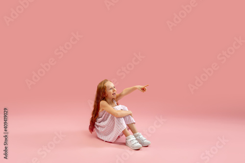 Sitting, pointing. Childhood and dream about big and famous future. Pretty longhair girl on coral pink studio background. Childhood, dreams, imagination, education, facial expression, emotions concept