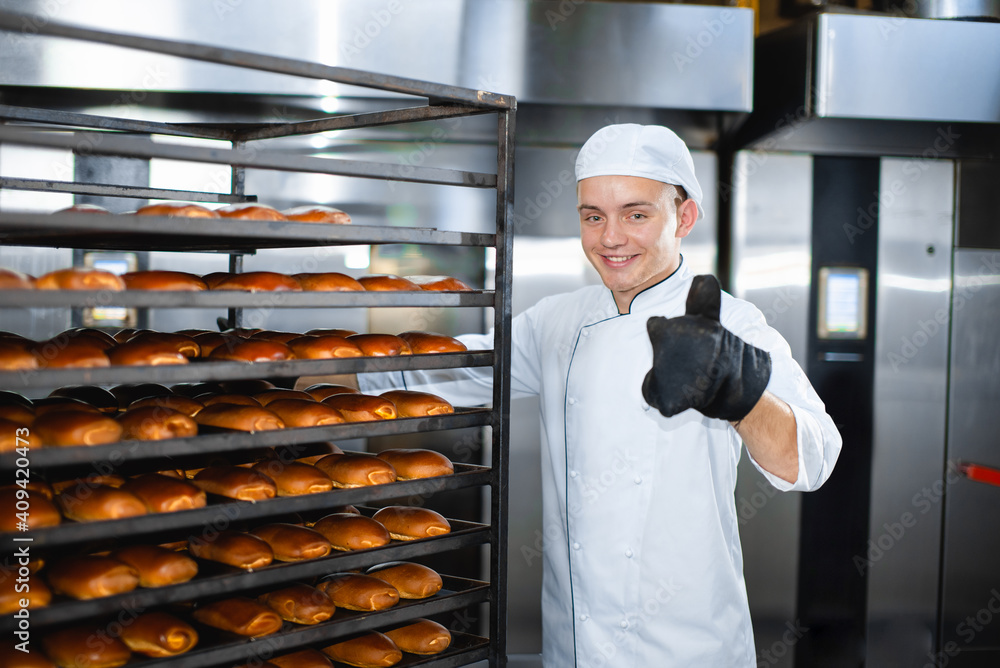 Obraz na plátně Baker with a hot baking trolley on a background of an industrial oven shows thumb up