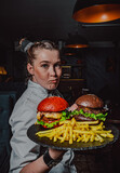 young woman chef with two burger on dark background in cafe
