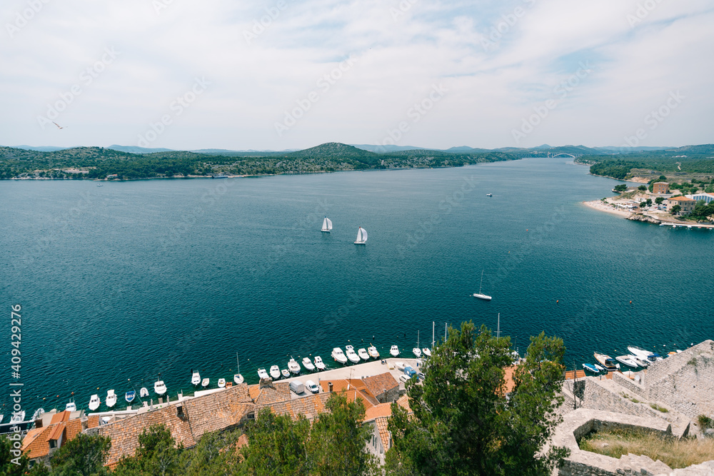 View of the embankment with moored boats and two sailing ships sailing in the sea from the walls of the old town of Sibennik in Croatia.