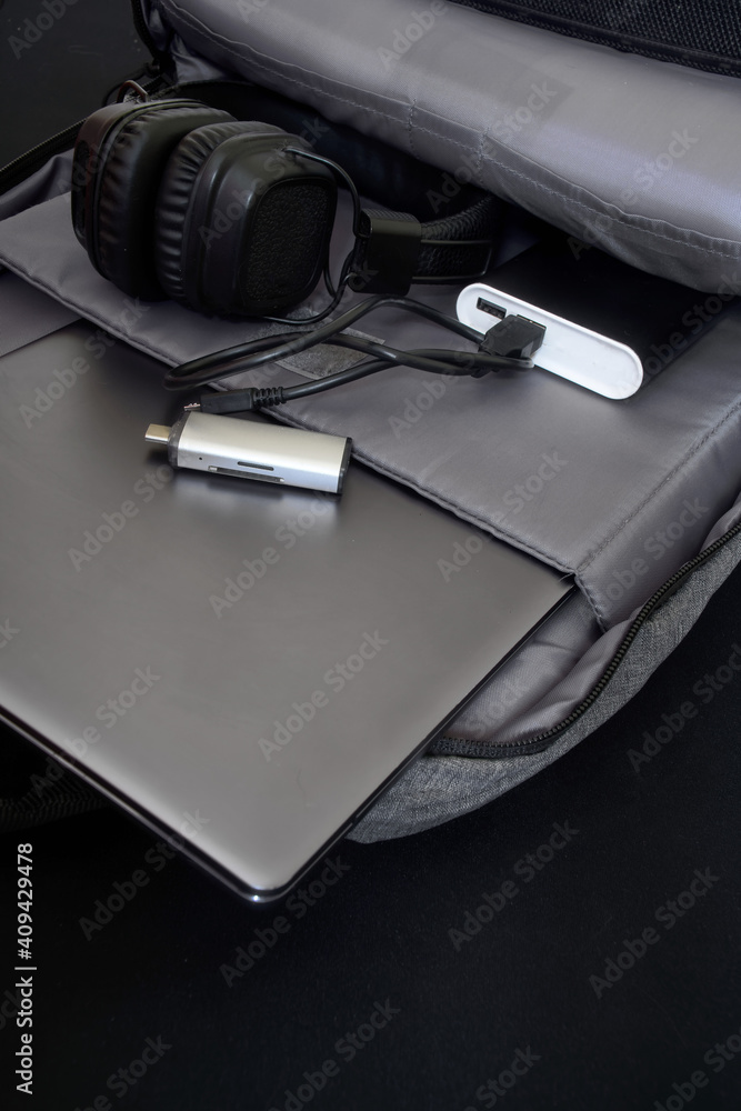 An open gray urban backpack containing a USB card reader, power bank, wireless headphones, and a silver laptop or ultrabook. An open gray urban backpack containing a USB card reader, power bank, wireless headphones, and a silver laptop or ultrabook.
