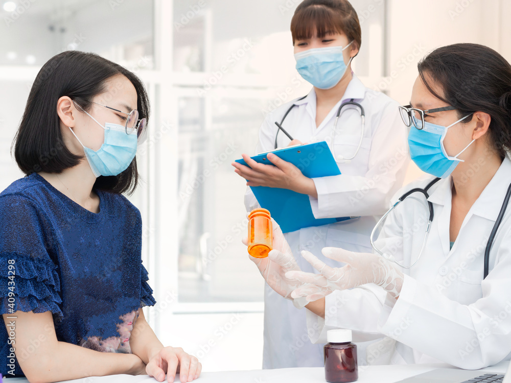 Female doctor holding pill bottle explaining how to take medicine to cure the virus or flu disease to a woman in hospital, trainee is standing beside taking note on a chart Female doctor holding pill bottle explaining how to take medicine to cure the virus or flu disease to a woman in hospital, trainee is standing beside taking note on a chart