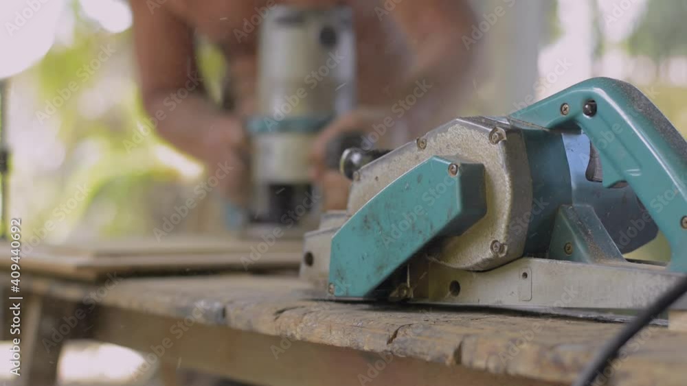 Blurred carpenter use professional tools for crafting. Close up electric planer with old male carpenter working with electric router on wooden slats. DIY maker and carpentry work concept. Blurred carpenter use professional tools for crafting. Close up electric planer with old male carpenter working with electric router on wooden slats. DIY maker and carpentry work concept.
