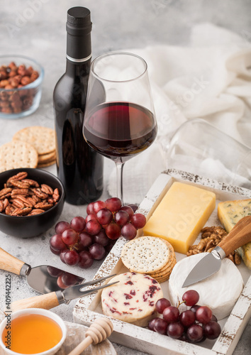 Bottle and glass of red wine with selection of various cheese in wooden box and grapes on light table background. Blue Stilton, Red Leicester and Brie Cheese with Cheddar and nuts with honey.