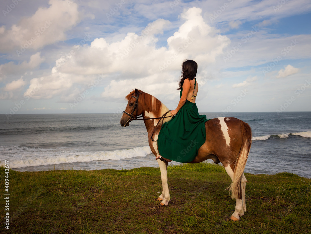 Woman riding horse near the ocean. Outdoor activities. Asia woman wearing long green dress. Traveling concept. Cloudy sky. View from back. Copy space. Bali Woman riding horse near the ocean. Outdoor activities. Asia woman wearing long green dress. Traveling concept. Cloudy sky. View from back. Copy space. Bali