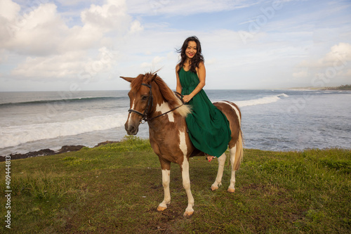 Happy woman riding horse near the ocean. Outdoor activities. Asia woman wearing long green dress. Traveling concept. Cloudy sky. Copy space. Bali
