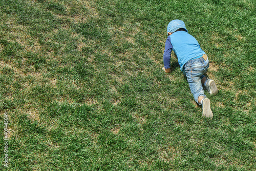 Playful little boy in blue cap goes up steep hill slope covered with lush green grass in spring park on sunny day backside view