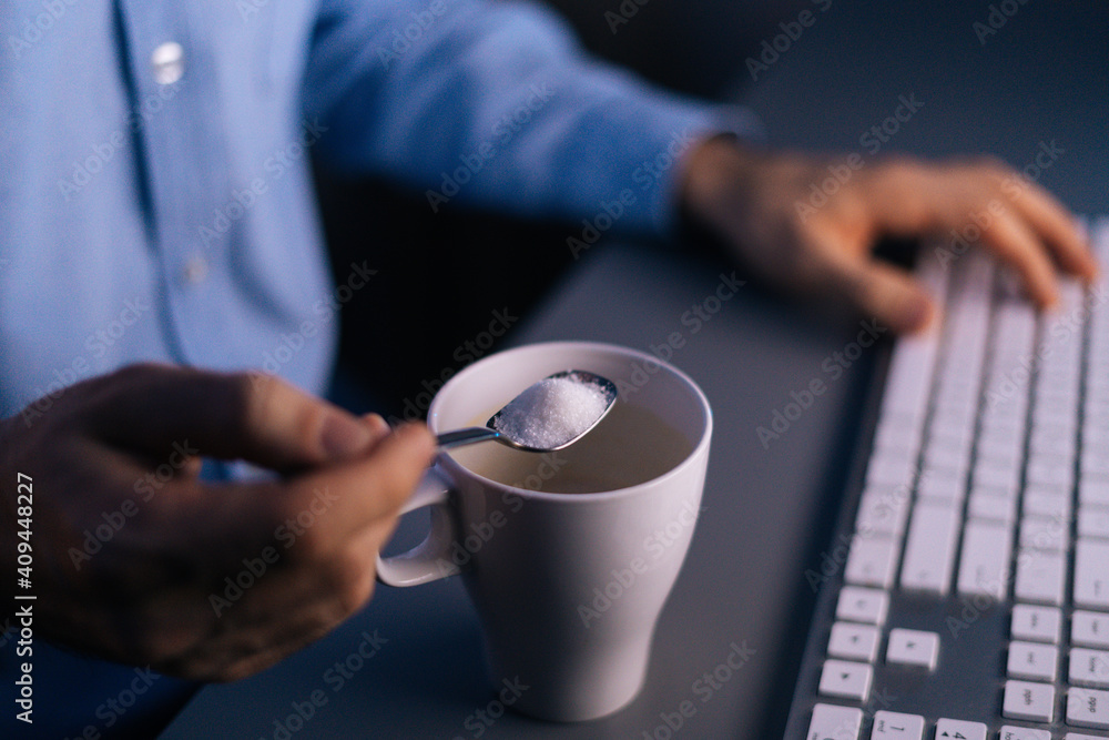 Close-up view of hands unrecognizable man pouring sugar on coffee in cup with small metal spoon during working on computer at late night. Concept of remote working, distance learning. 