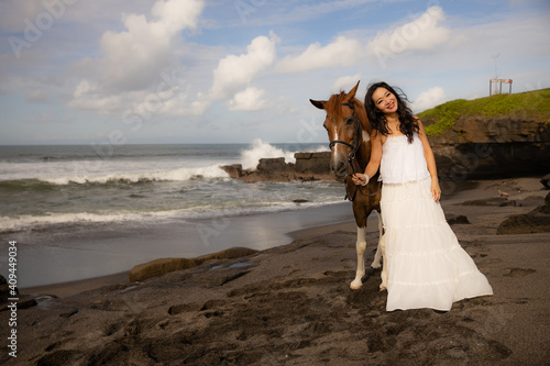 Smiling woman leading horse by its reins. Horse riding on the beach. Human and animals relationship. Asian woman wearing long white dress. Bali, Indonesia