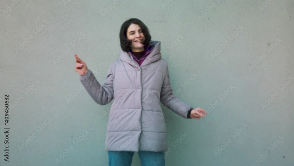 Front view portrait of young beautiful woman standing against a grey wall, looking at the camera, dancing and expressing happiness. Front view portrait of young beautiful woman standing against a grey wall, looking at the camera, dancing and expressing happiness.