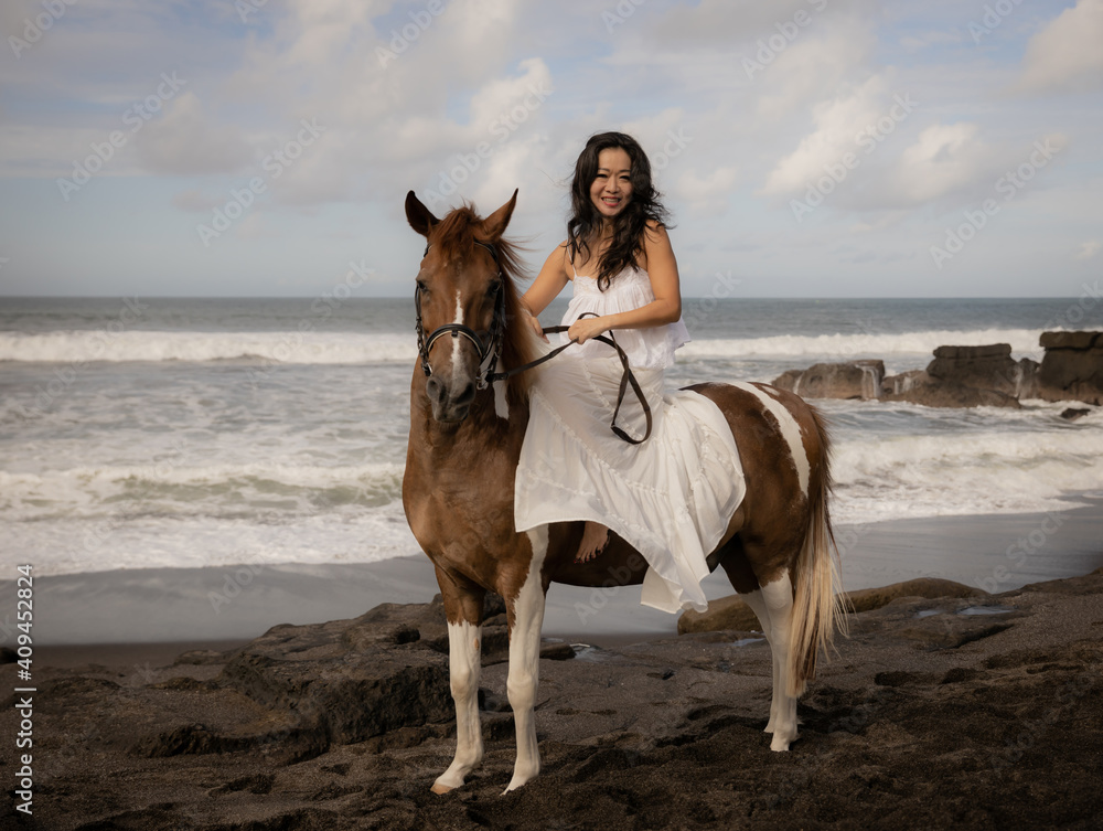 Asian woman riding horse on the beach. Outdoor activities. Woman wearing long white dress. Traveling concept. Cloudy sky. Copy space. Bali Asian woman riding horse on the beach. Outdoor activities. Woman wearing long white dress. Traveling concept. Cloudy sky. Copy space. Bali