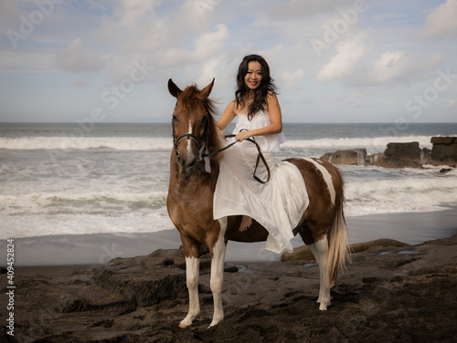 Asian woman riding horse on the beach. Outdoor activities. Woman wearing long white dress. Traveling concept. Cloudy sky. Copy space. Bali