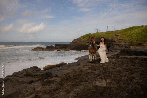 Woman leading horse by its reins. Horse riding on the beach. Human and animals relationship. Asian woman wearing long white dress. Nature concept. Copy space. Bali