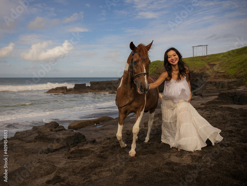 Happy woman leading horse by its reins. Horse riding on the beach. Human and animals relationship. Asian woman wearing long white dress. Nature concept. Copy space. Bali, Indonesia