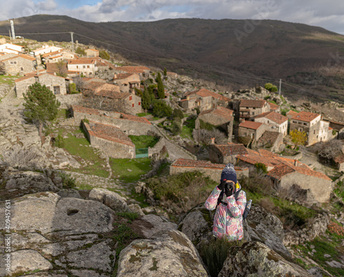 Photo of a young and attractive female standing on top of a rock and taking photos with the village of Trevejo at the back in the north of Spain. photo