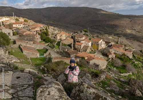 Photo of a young and attractive female standing on top of a rock and taking photos with the village of Trevejo at the back in the north of Spain. photo