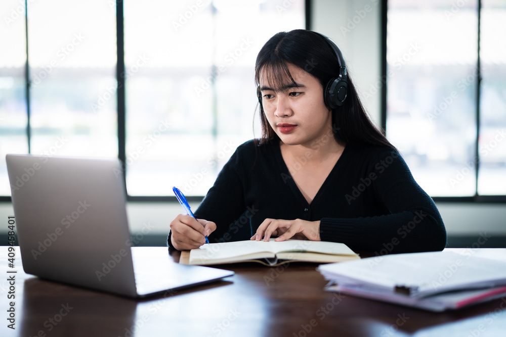 Smiling Asian girl student wears wireless headphones write on the notebook to study language online watch and listen to the lecturer, webinar via video call e-learning at home, distance education