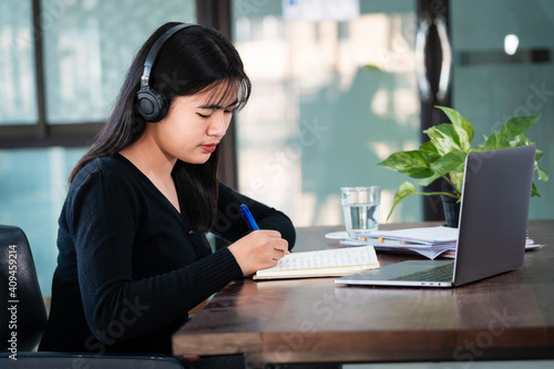 Smiling Asian girl student wears wireless headphones write on the notebook to study language online watch and listen to the lecturer, webinar via video call e-learning at home, distance education