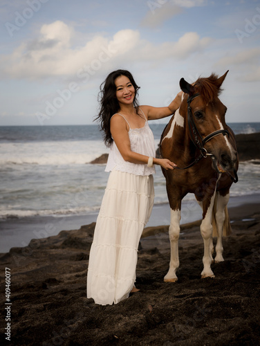 Woman leading horse by its reins. Horse riding on the beach. Human and animals relationship. Asian woman wearing long white dress. Travel concept. Copy space. Bali