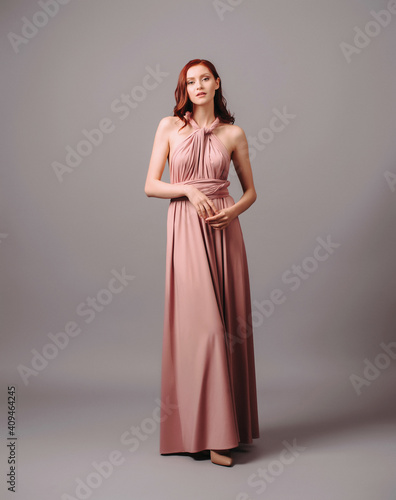 Studio portrait of pensive ginger glamour young woman in pink halter dress on grey background. Bridesmaid's look. Natural makeup and hairdo.