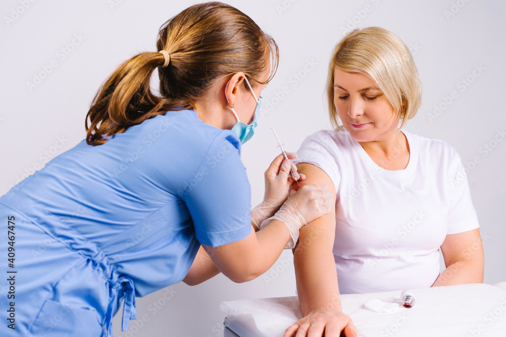 A doctor in blue medical uniform puts the vaccine with a syringe to a senior blonde woman. White background. A doctor in blue medical uniform puts the vaccine with a syringe to a senior blonde woman. White background.