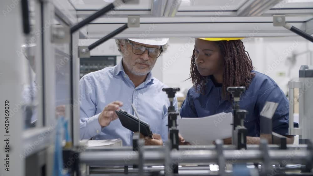 Older factory engineer training black employee to operate new industrial machine, using tablet and paper manual. Production process or machinery concept